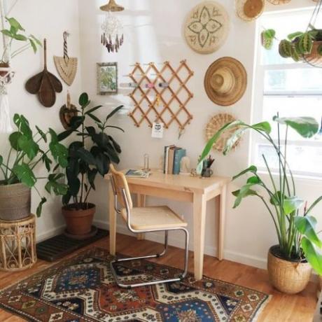 „Boho“ namų biuro kampas su patraukliu baltu stalu, rotango kėde, „macrame“, dideliais kambariniais augalais.